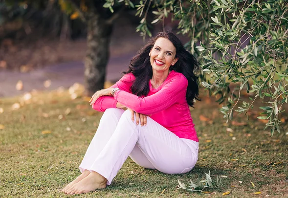 A woman in a pink top and white pants sits barefoot on grass near a leafy tree, smiling.