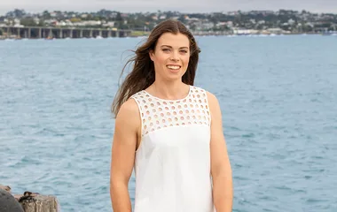 A woman in a white dress stands smiling by a body of water with a distant shoreline in the background.