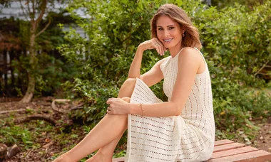 A woman in a light dress sits on a bench in a garden, smiling with her chin resting on her hand.