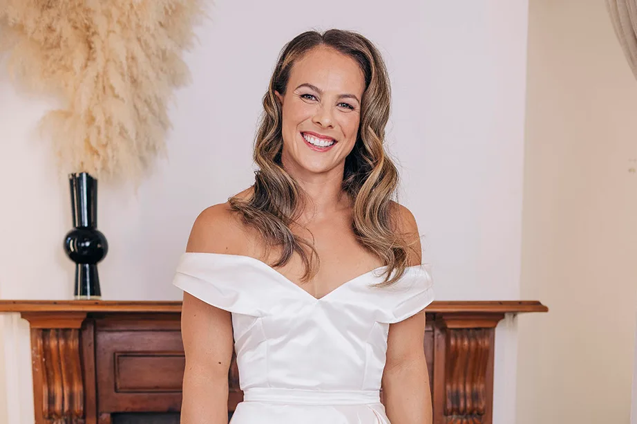 A woman in a white off-shoulder dress smiling in a room with a wooden fireplace and decorative pampas grass.