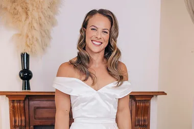 A woman in a white off-shoulder dress smiling in a room with a wooden fireplace and decorative pampas grass.