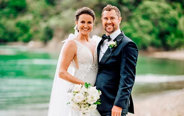 Bride in lace gown and groom in tuxedo smiling on a beach, holding a bouquet of white flowers.