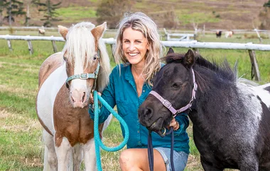 Smiling woman kneeling with two small horses on a grassy field.