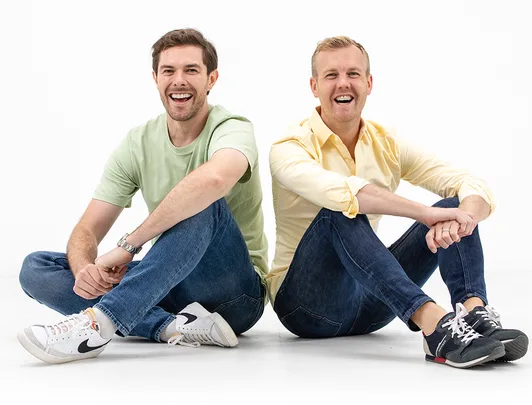 Two men sitting on the floor, smiling. One in a green shirt and white sneakers, the other in yellow with black sneakers.