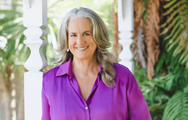 A woman with long gray hair smiles in a purple shirt, standing outdoors near a white column and greenery.