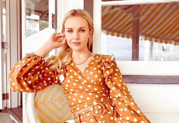 Woman in an orange polka dot dress sitting on a chair indoors, with yellow-striped awning outside.