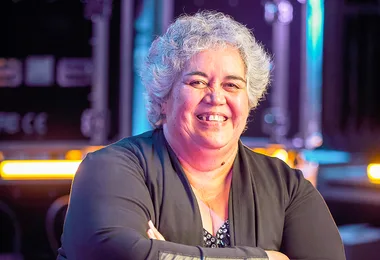 A smiling person with short gray hair in a black outfit stands in a dimly lit area with soft focus, colorful background lights.