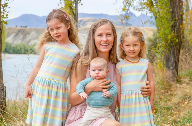 Mother with three children, two girls in striped dresses and a baby, pose by a river under trees with hills in the background.