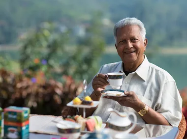 Senior man smiling and holding a teacup outdoors with a plate of treats on the table.