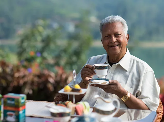Senior man smiling and holding a teacup outdoors with a plate of treats on the table.