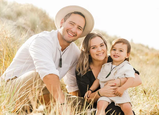 A happy family of three sits together in a grassy field, all smiling at the camera.