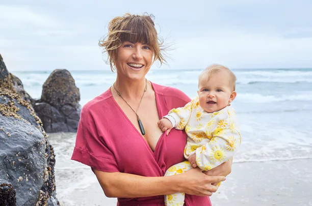 ```
Woman holding a smiling baby on a beach with rocks and waves in the background.
```