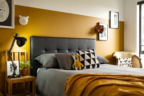 Cozy bedroom with mustard accent wall, black lamp, and decorative pillows on a gray bed, near a window with natural light.