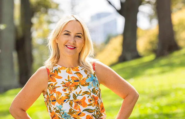 Blonde woman in floral dress smiling in a sunny park setting.