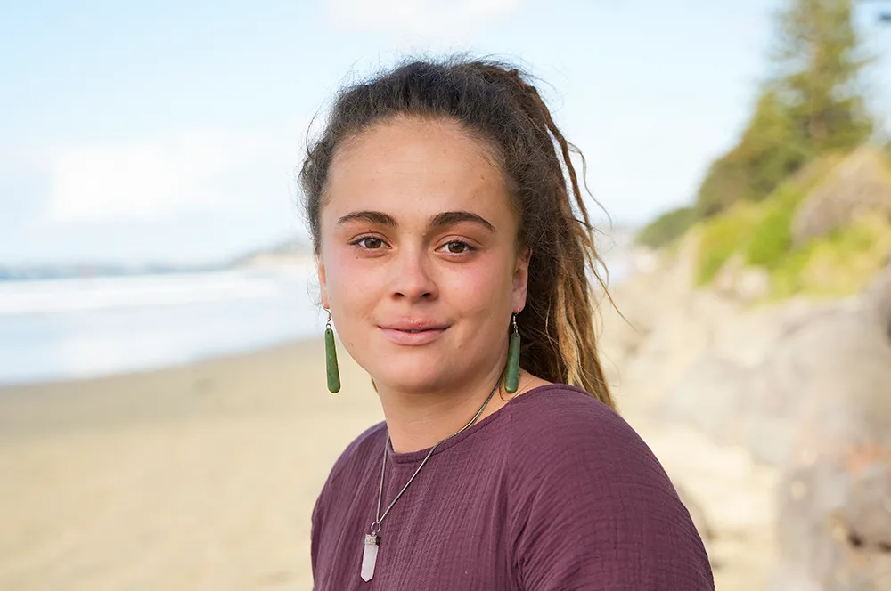 Young woman with long hair and jade earrings stands on a beach in natural light, smiling softly at the camera.