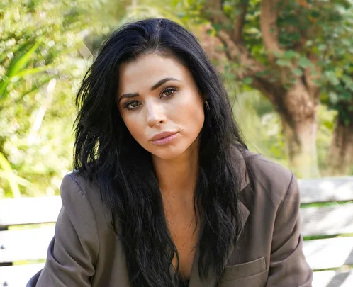 Woman with long black hair in a blazer, sitting on a bench in a green park setting, looking at the camera.