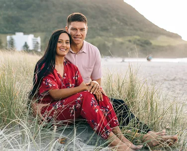 Young couple sitting on a sandy beach with grassy dunes, mountain and buildings in the background.
