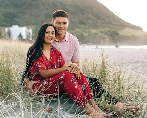 Young couple sitting on a sandy beach with grassy dunes, mountain and buildings in the background.