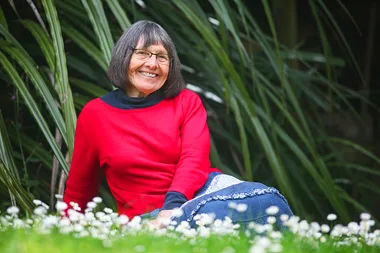 A smiling woman in a red sweater sits on grass with white flowers, in front of tall green plants.