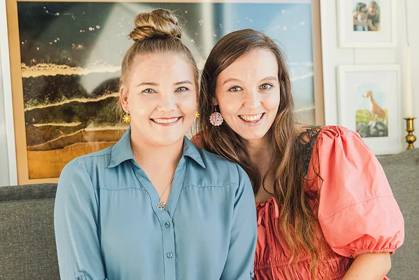 Two women smiling, sitting closely on a couch with framed artwork in the background.