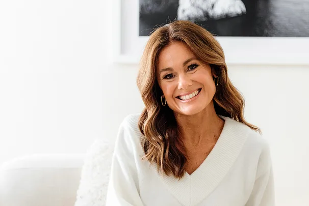 A woman with brown hair smiling, wearing a white sweater, sitting indoors with a framed picture in the background.