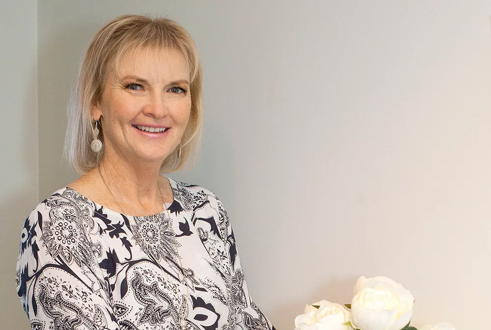 A woman smiling, wearing a patterned top, with short blonde hair, standing near white flowers.