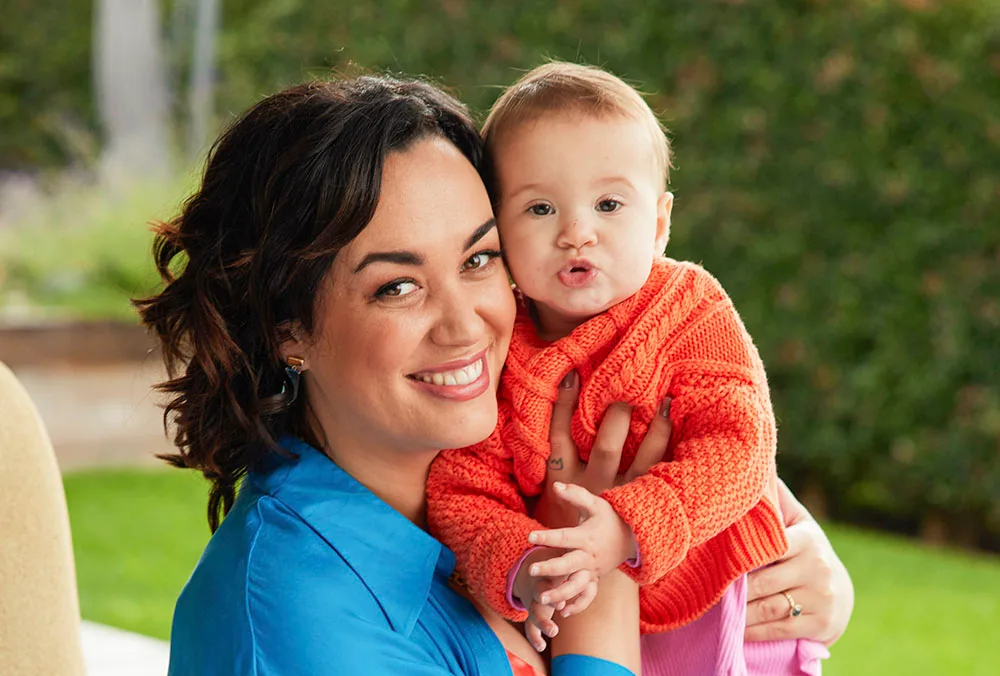 Woman in blue shirt holding a baby in an orange sweater, both smiling outdoors.