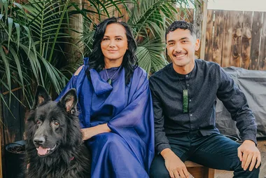 Two people sitting beside a black dog, with green plants in the background, both wearing dark clothing.