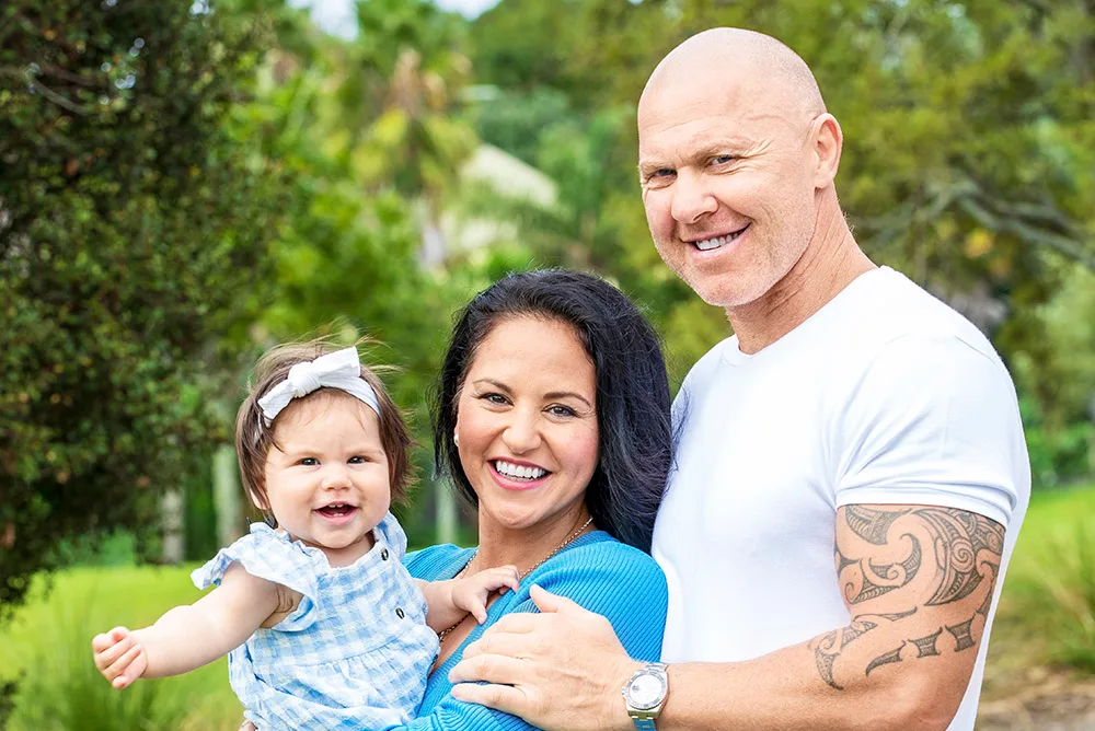 Happy family posing outdoors, woman holding a baby, with a smiling man standing beside them in a lush green setting.
