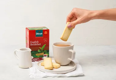 Hand dipping biscuit into tea with Dilmah English Breakfast tea box and small jug nearby.