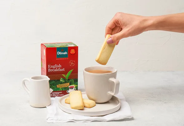 Hand dipping biscuit into tea with Dilmah English Breakfast tea box and small jug nearby.