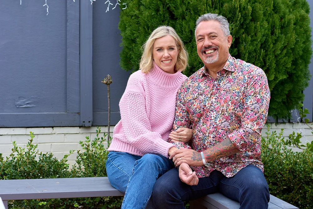 A smiling couple sitting on a bench, the woman in a pink sweater and man in a floral shirt, with greenery in the background.