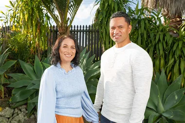 A man and woman smiling, standing outdoors with tropical plants in the background.