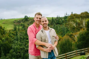A couple smiling and embracing outdoors, with a lush green hillside in the background.