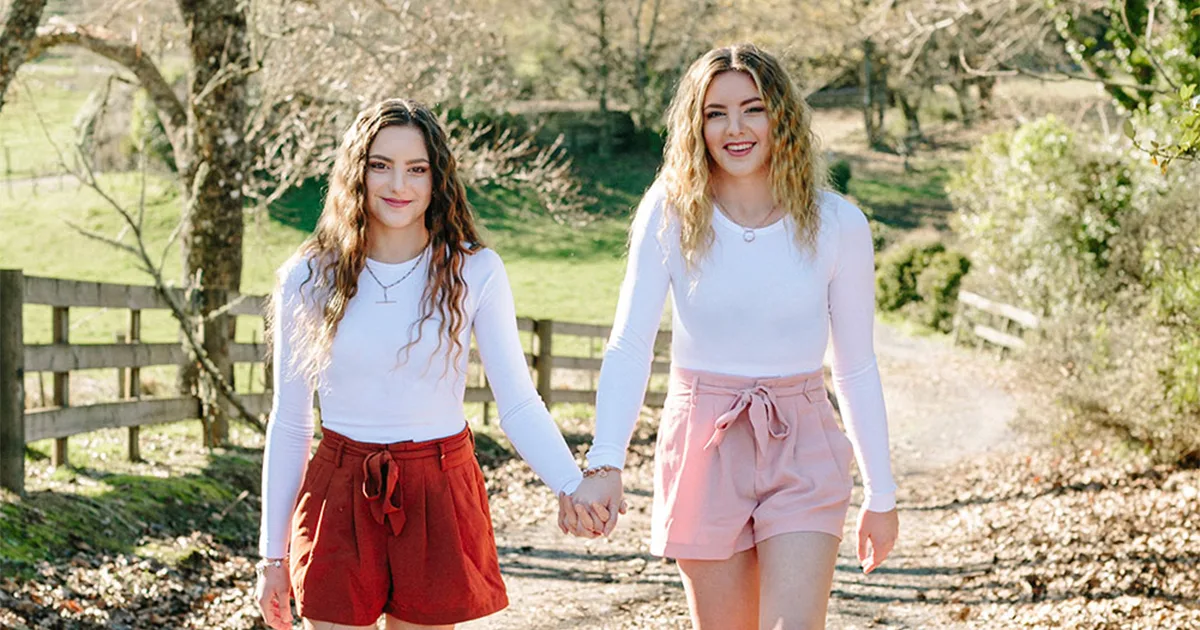 Two young women walking hand in hand outdoors, both in white tops and red shorts, smiling.