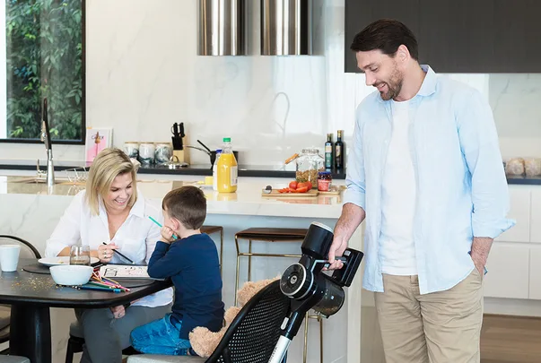 Family in a kitchen: woman drawing with boy at table, man holding a vacuum cleaner, kitchen items in background.