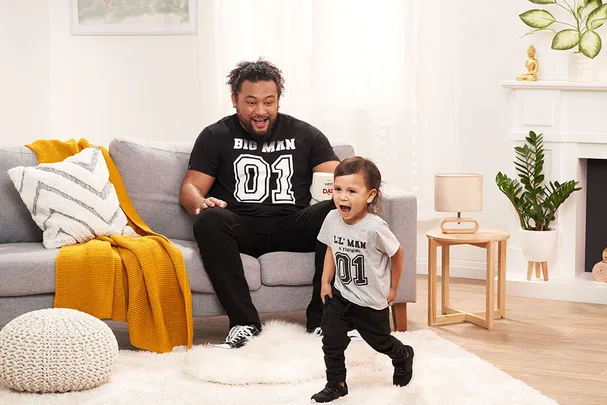 Father and child in matching outfits, sitting on a couch in a cozy living room, smiling and interacting playfully.