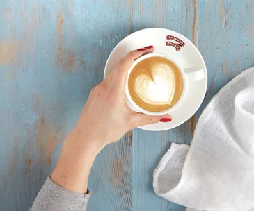 A hand with red nails holds a cappuccino with heart latte art on a blue wooden table beside a white napkin.
