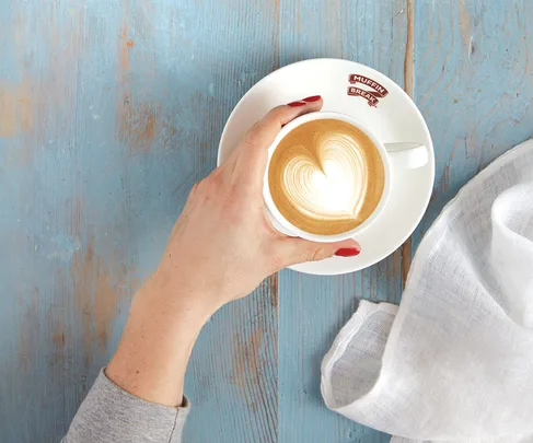 A hand with red nails holds a cappuccino with heart latte art on a blue wooden table beside a white napkin.