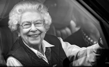 Woman with curly hair and glasses smiling while sitting in a car, photographed in black and white.