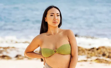 A woman in a green bikini stands on a beach with ocean waves in the background.