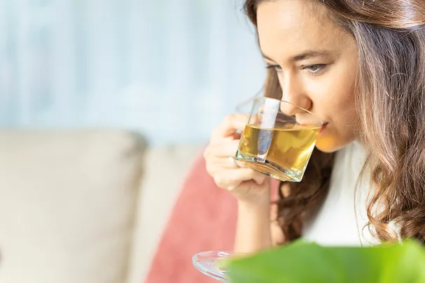 A woman sips green tea from a clear glass cup while sitting on a sofa.