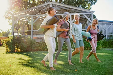 Four women walking barefoot on grass with yoga mats and water bottles, laughing and chatting in a sunny park setting.