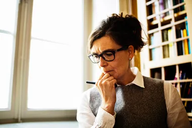 A woman in glasses, deep in thought, holds a pen to her lips in a book-filled room by a bright window.