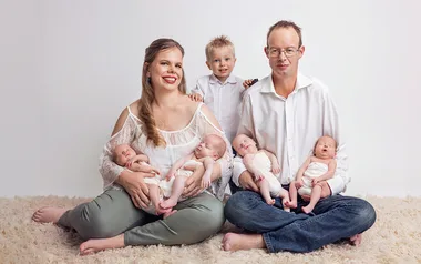 Family portrait with a smiling woman and man sitting cross-legged, each holding two babies, and a child standing behind them.
