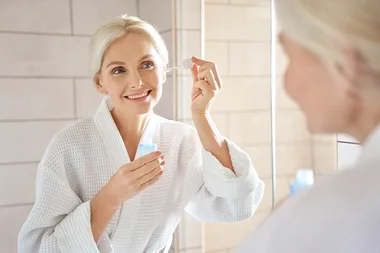 An older woman in a white robe applies serum with a dropper while smiling in front of a bathroom mirror.
