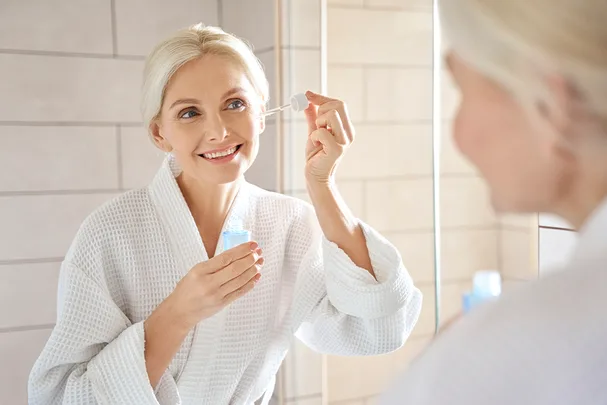 An older woman in a white robe applies serum with a dropper while smiling in front of a bathroom mirror.