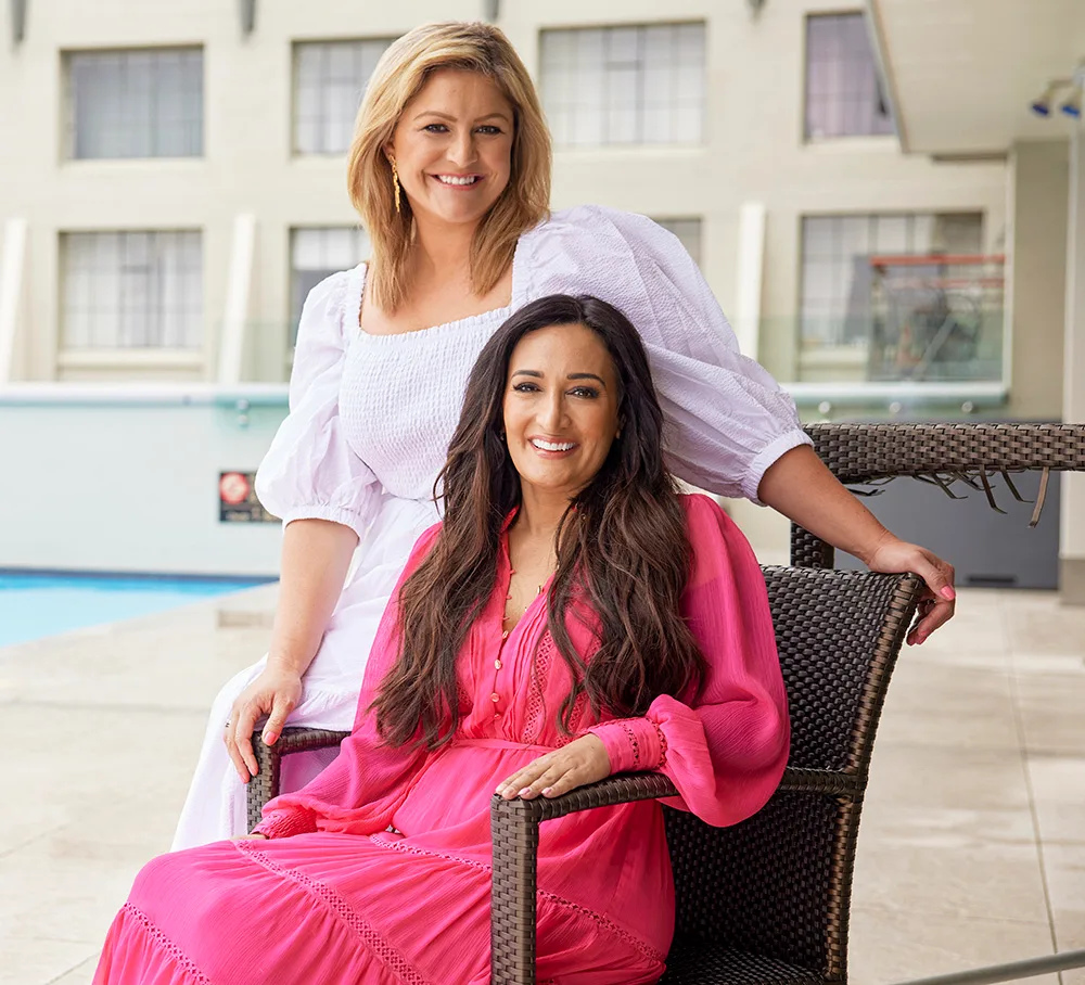 Two women smiling by a pool; one seated wearing pink, the other standing in white, with a modern building backdrop.