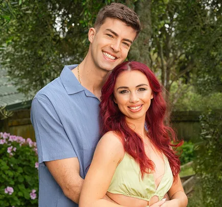 Couple smiling outdoors, man in blue shirt, woman with red hair in light green outfit, surrounded by greenery.