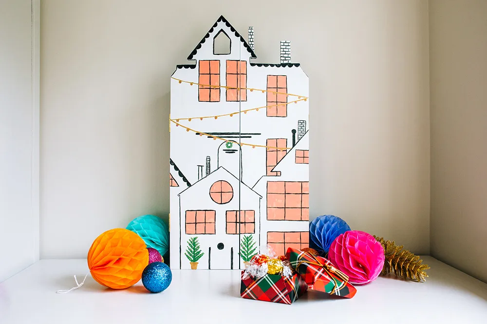 Cardboard house with drawn windows and lights, surrounded by colorful paper balls and wrapped gifts.